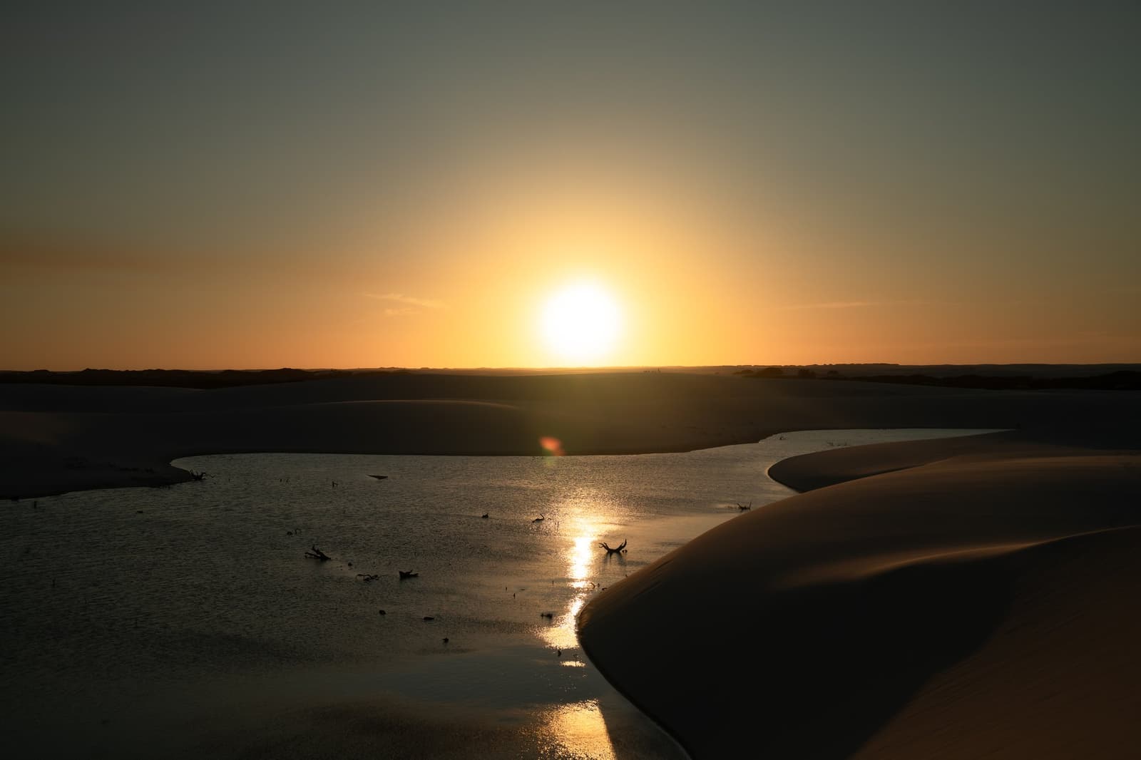 The hour when the dunes decide to turn gold and the birds couldn't care less. Sun setting on the horizon over a shallow lagoon nestled between dark sand dunes on a Lençóis Maranhenses trip, with birds silhouetted on the glassy water