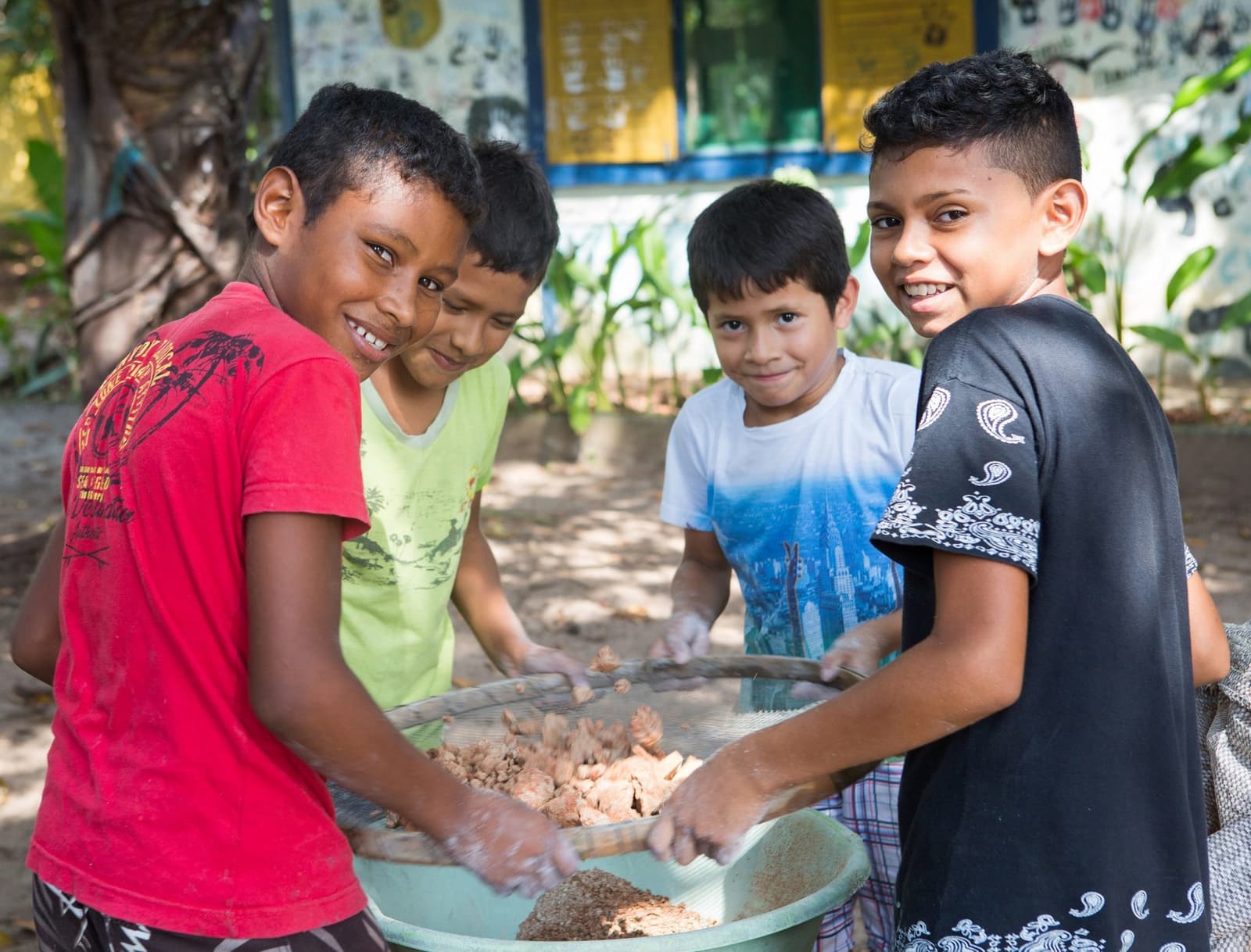 Four pairs of hands, one shared task, and plenty of laughter. Four smiling boys work together sifting and sorting food in a large basin outdoors, part of a community engagement activity at a luxury amazon lodge brazil experience