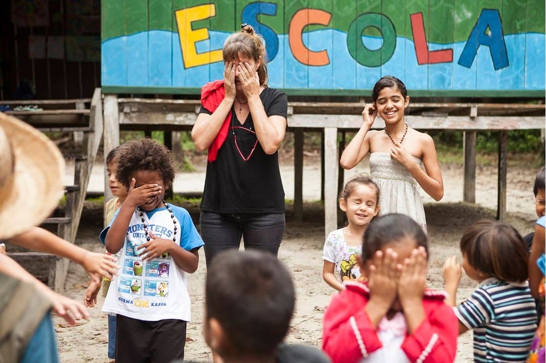 The rules of the game? Nobody told her. The kids know exactly what they're doing. A traveller and local children play a hand-covering game in front of a colourfully painted school building marked 'ESCOLA' during a community visit on a luxury Amazon lodge Brazil itinerary