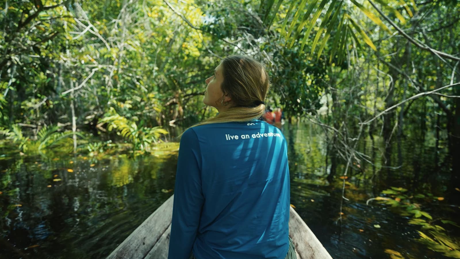 The forest doesn't open up, it pulls you in, one quiet paddle at a time. A traveller seated at the bow of a wooden canoe gazes up at the dense flooded forest canopy during a luxury Amazon lodge Brazil boat excursion through dark mirror-still waters