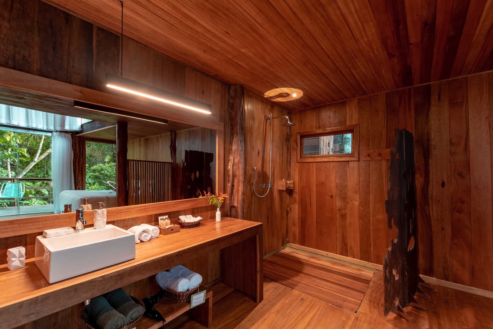Hot water, thick towels, and the forest just outside the glass. Warmly lit ensuite bathroom at a luxury amazon lodge brazil, featuring floor-to-ceiling certified hardwood panelling, a vessel sink vanity, rainfall shower, and a large mirror reflecting lush jungle greenery outside