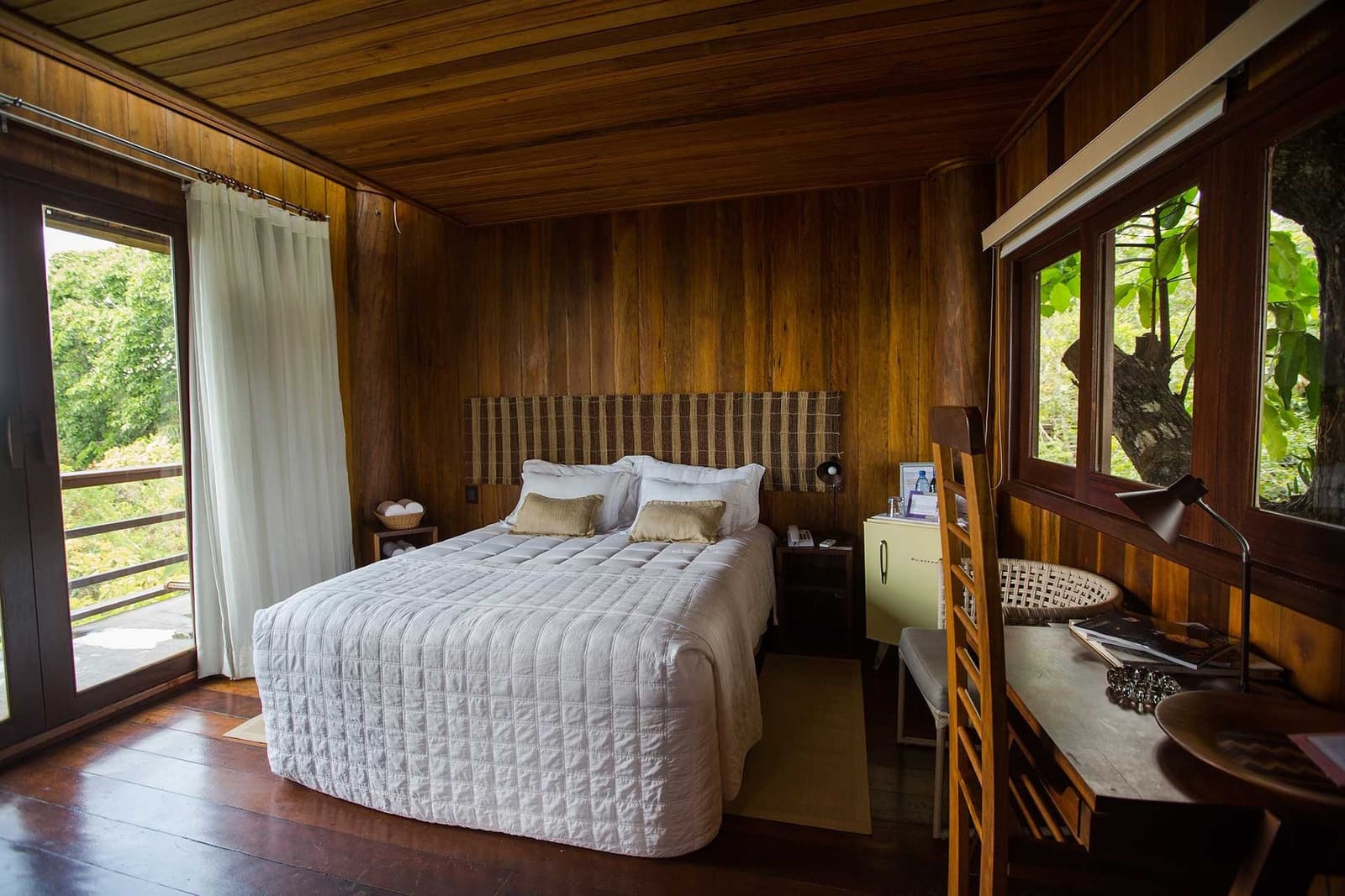 Sleeping this close to the canopy, mornings feel like a personal briefing from the forest. Cosy hardwood-panelled bedroom at a luxury amazon lodge brazil with a white quilted double bed, woven headboard, timber desk and lush green forest views through large windows