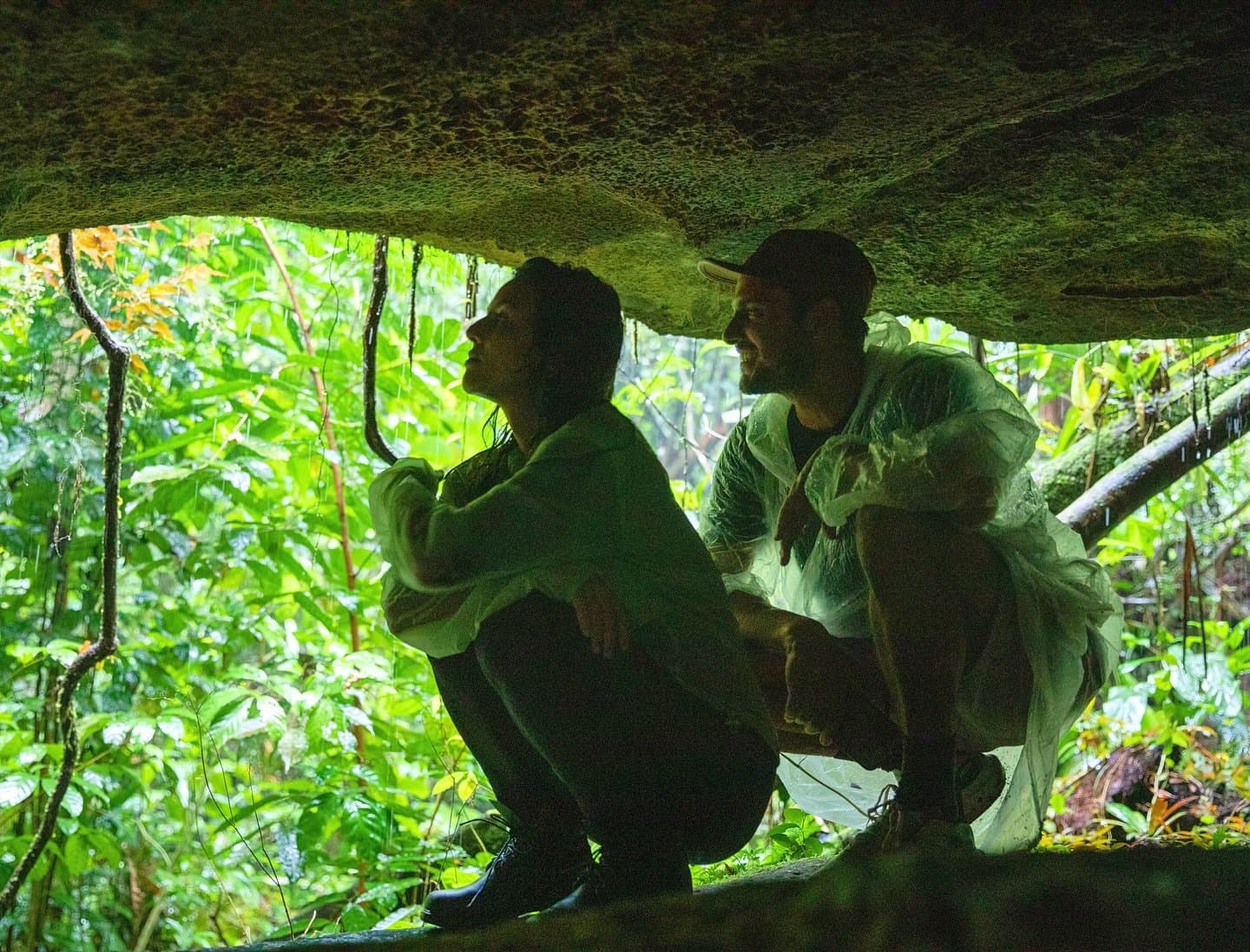 The forest finds ways to surprise you, even from underneath. Two travellers crouch beneath a mossy rock overhang at a luxury amazon lodge brazil cave trail, surrounded by dense green rainforest vegetation