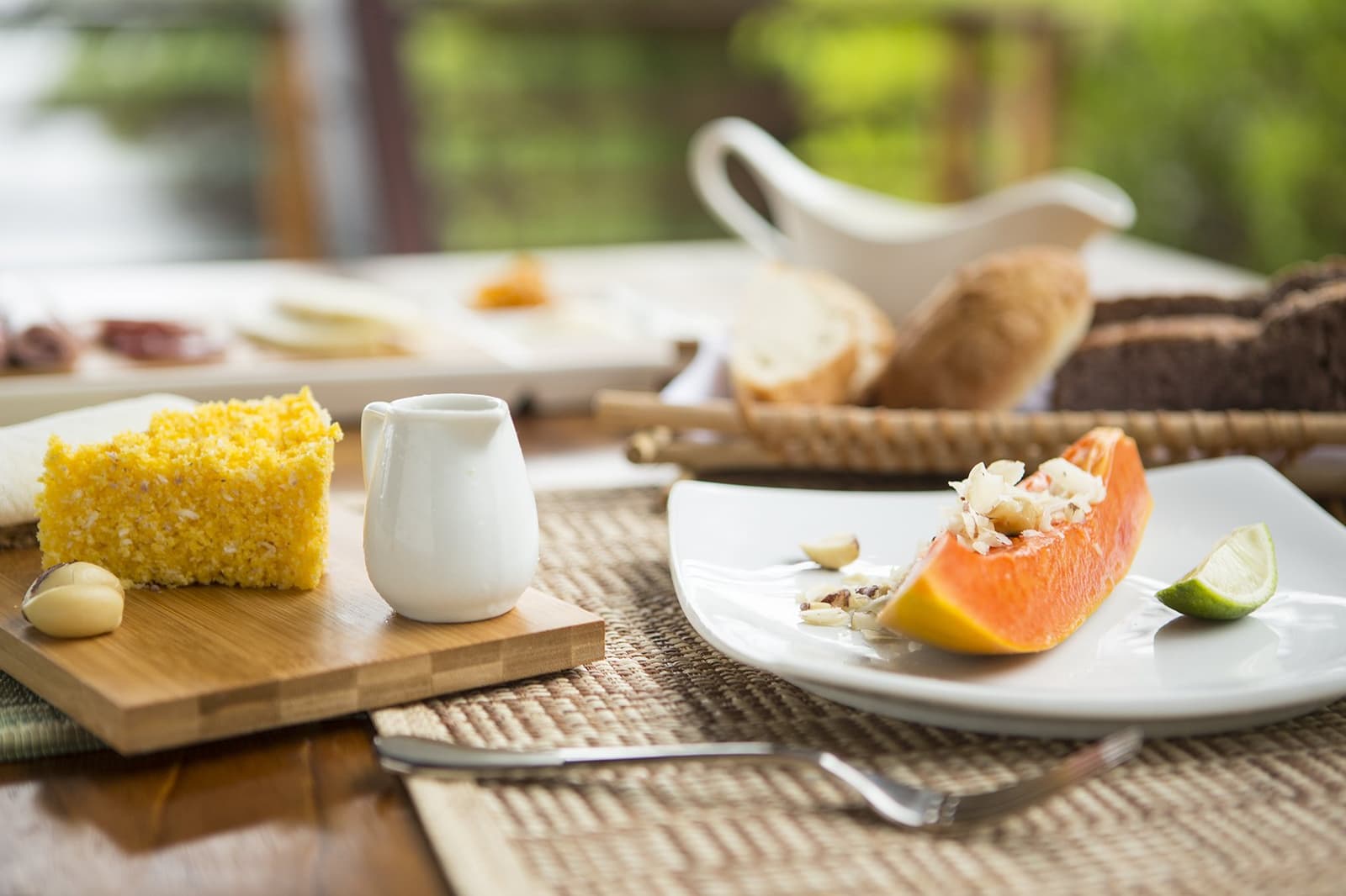 Breakfast here earns its place on the itinerary. Fresh papaya topped with nuts and lime, served alongside a slice of yellow corn cake on a bamboo board, part of a luxury Amazon lodge Brazil morning breakfast table set outdoors with lush greenery in the background