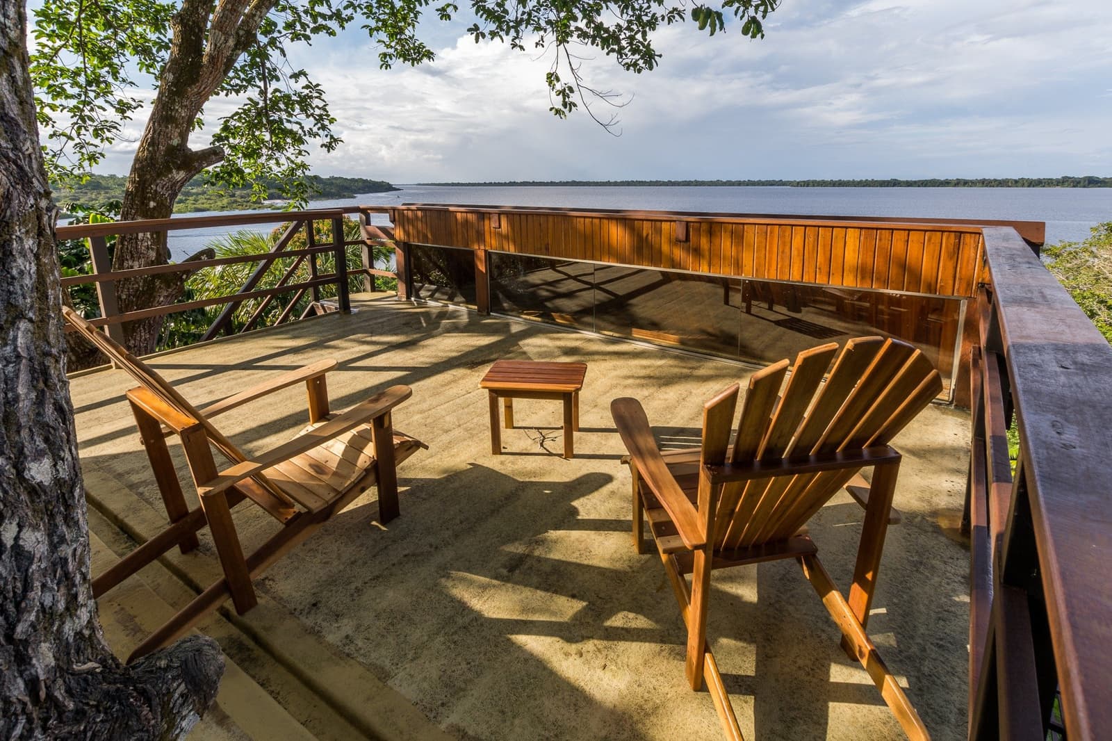 Your seat is ready, and the river isn't going anywhere. Two wooden Adirondack chairs and a small side table on an open hardwood deck at a luxury Amazon lodge Brazil, overlooking a wide dark river bordered by dense rainforest under a cloudy sky