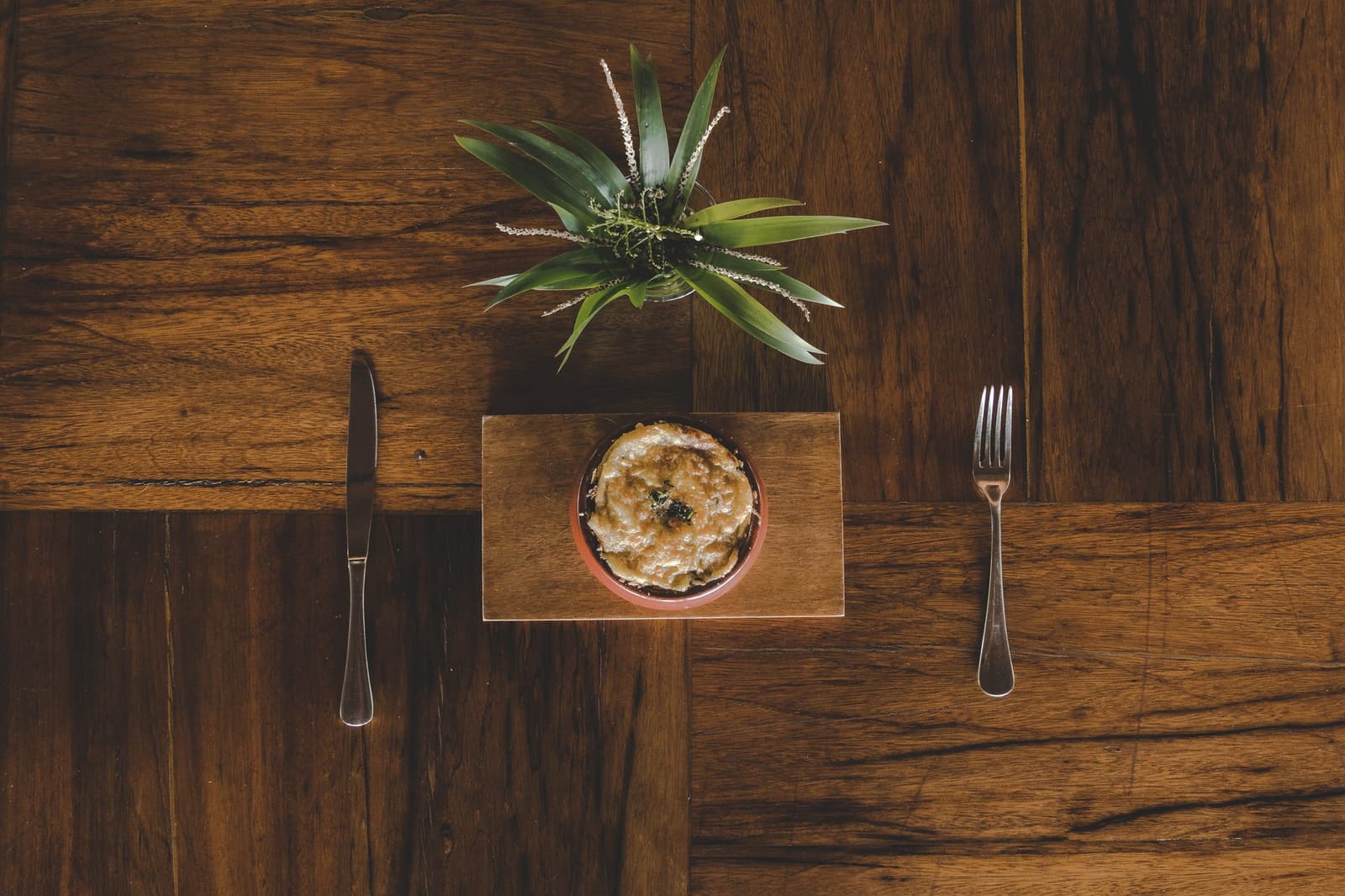 Good food deserves a good table, and the Amazon delivers on both. Overhead flat-lay of a baked dish in a clay ramekin served on a wooden board with knife and fork, part of the luxury Amazon lodge Brazil dining experience