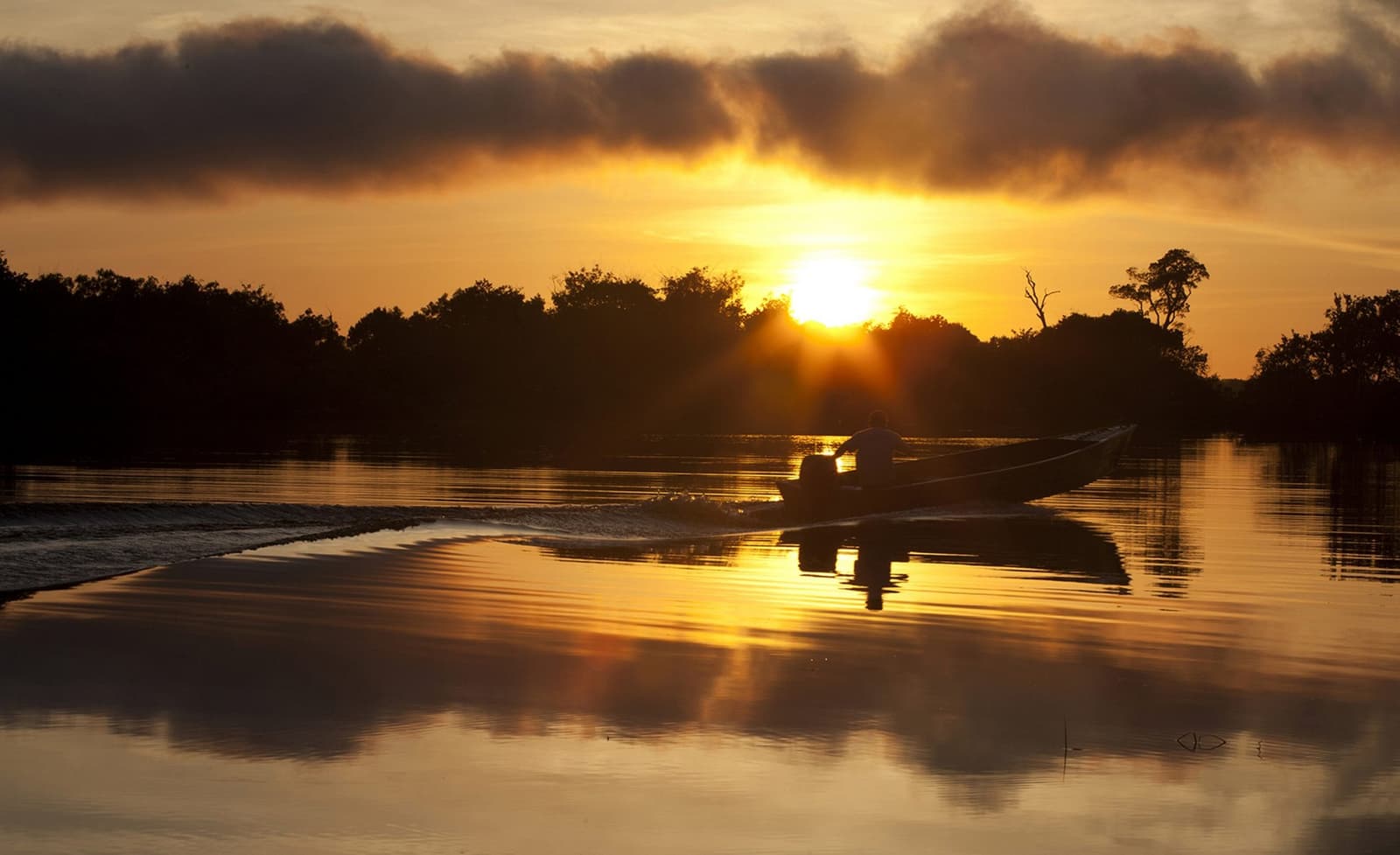 The river keeps its own schedule, and right now it says gold. A silhouetted motorboat glides across a glassy river toward a blazing golden sunset, with dark tree-lined banks reflected in the still water at a luxury Amazon lodge Brazil experience