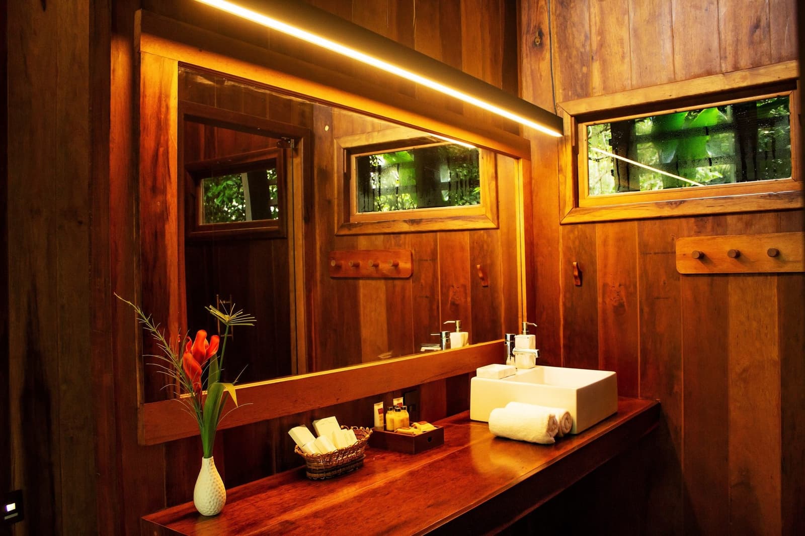 Jungle outside, warm wood within, the forest never felt so comfortable. Warmly lit en-suite bathroom at a luxury amazon lodge brazil, featuring polished hardwood walls, a white vessel sink, rolled towels, toiletry basket and a red tropical flower arrangement, with forest greenery visible through the windows