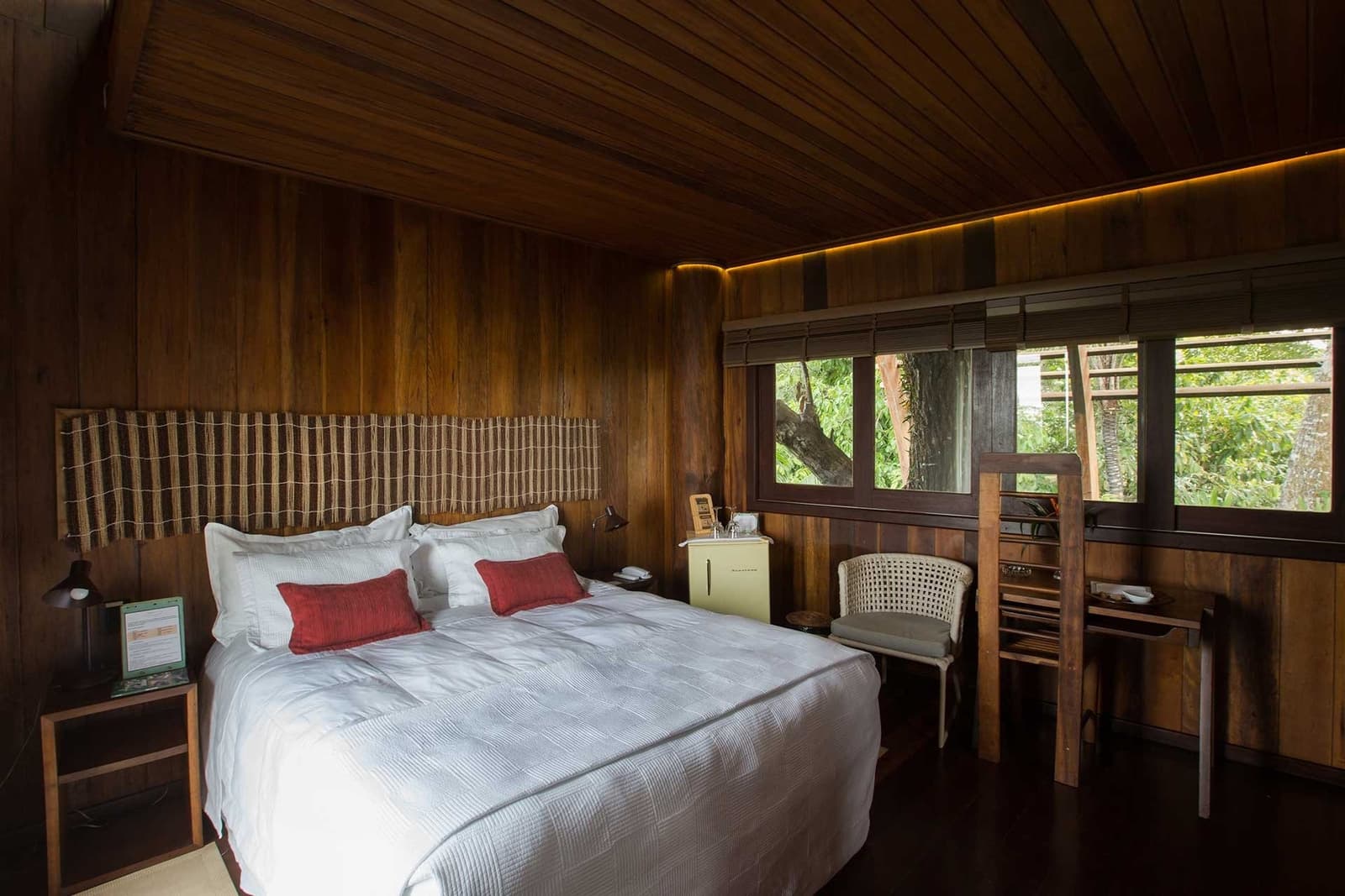 Fall asleep with the forest just inches from the glass. Warmly lit luxury amazon lodge brazil bedroom with dark hardwood walls and ceiling, a queen bed dressed in white linens and red accent cushions, and tall windows framing lush green rainforest canopy