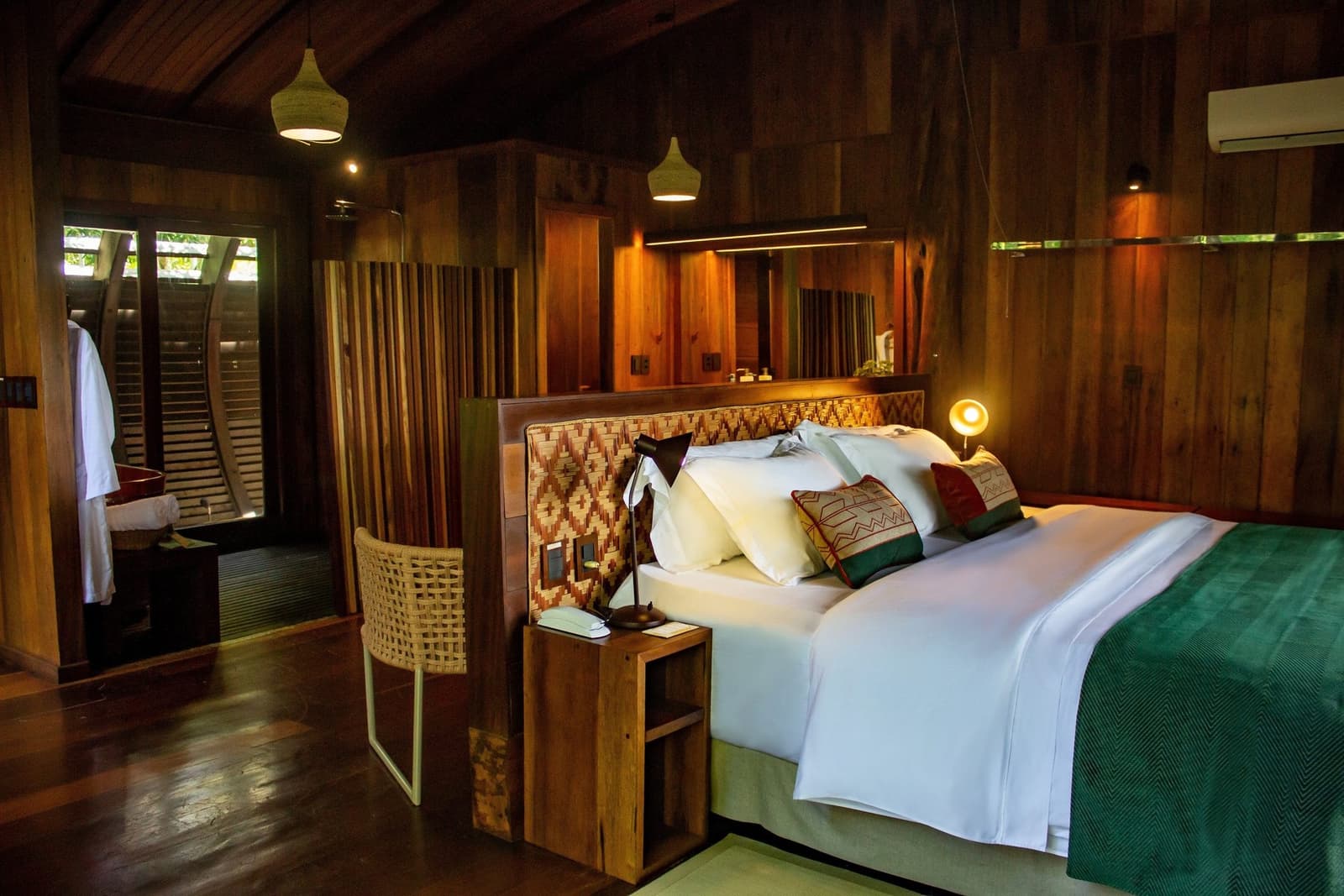 After a day on the river, this room earns every thread count. King bed with handwoven headboard and embroidered cushions inside a luxury Amazon lodge Brazil suite, surrounded by certified hardwood walls and warm pendant lighting
