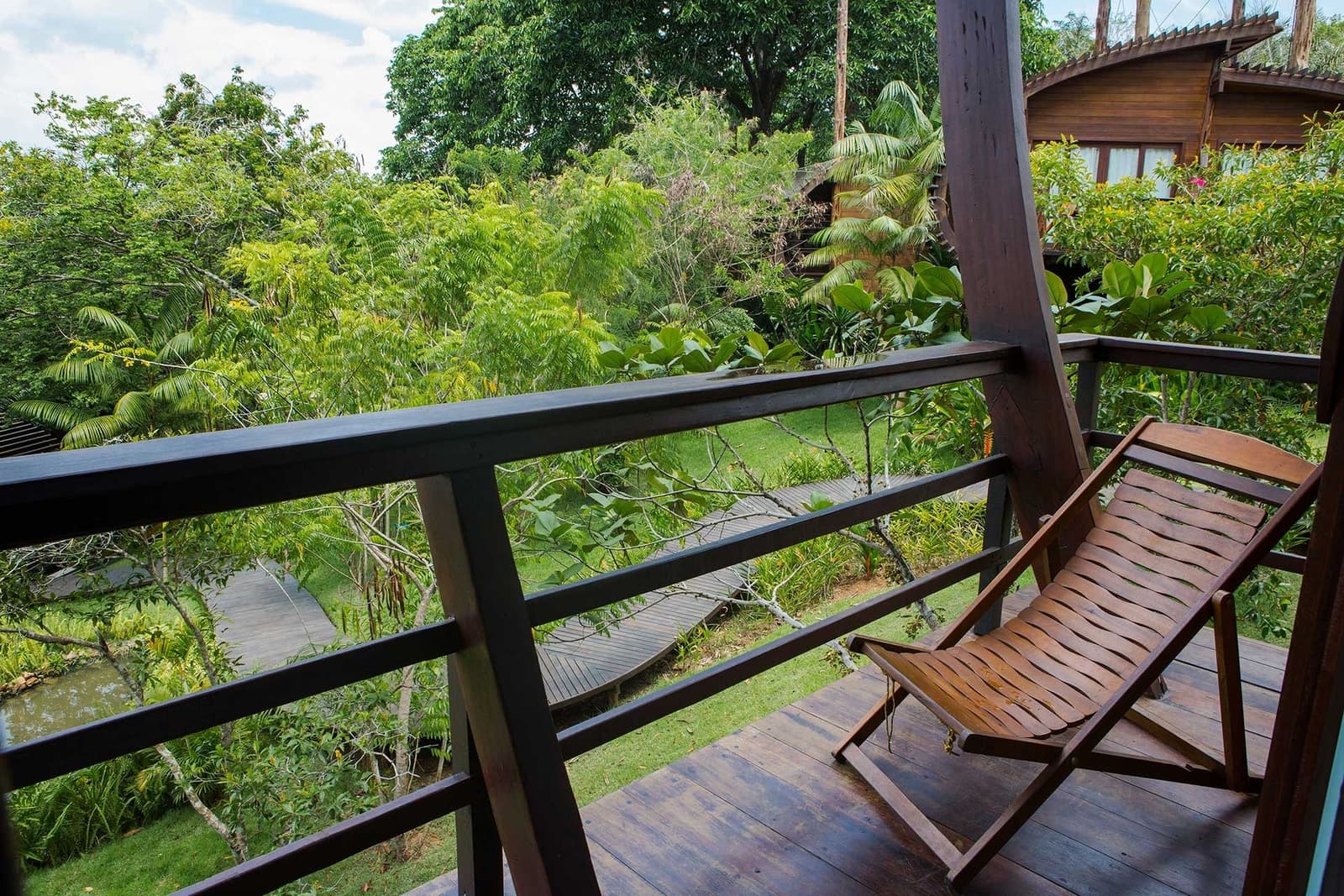 Your morning coffee spot, sorted, the garden does the rest. A wooden deck chair sits on a private hardwood balcony at a luxury amazon lodge brazil, overlooking a lush canopy, timber walkways, and a neighbouring bungalow nestled among tropical trees