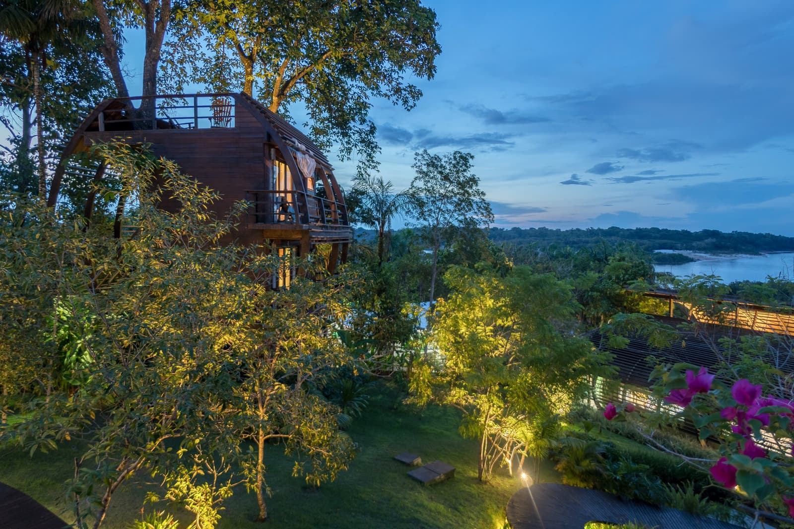 Your room for the night, suspended above the forest with a river view thrown in. A curved timber treehouse villa at a luxury amazon lodge brazil glows with warm interior light at dusk, surrounded by lush tropical garden with bougainvillea blooms and a wide river visible through the canopy in the background