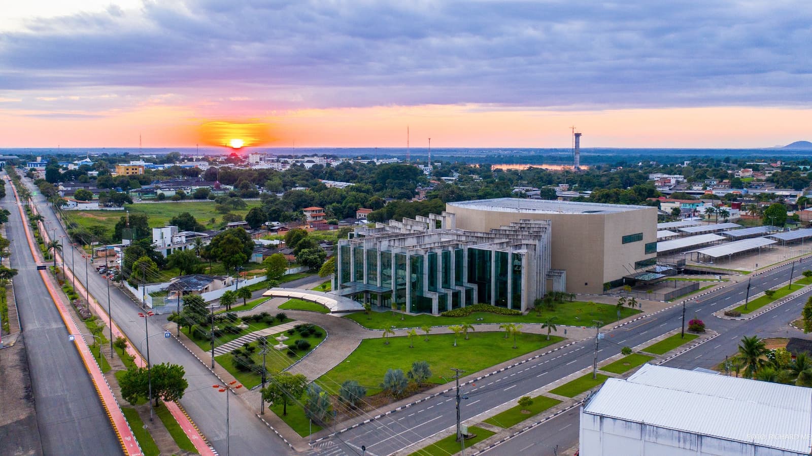 Your Roraima adventure starts at street level, before the tepuis even come into view. Aerial view of a Brazilian city at sunset, with a modernist glass-and-concrete civic building surrounded by green lawns and wide avenues, a glowing orange sun sinking toward the flat horizon — the gateway city for a Mount Roraima hike
