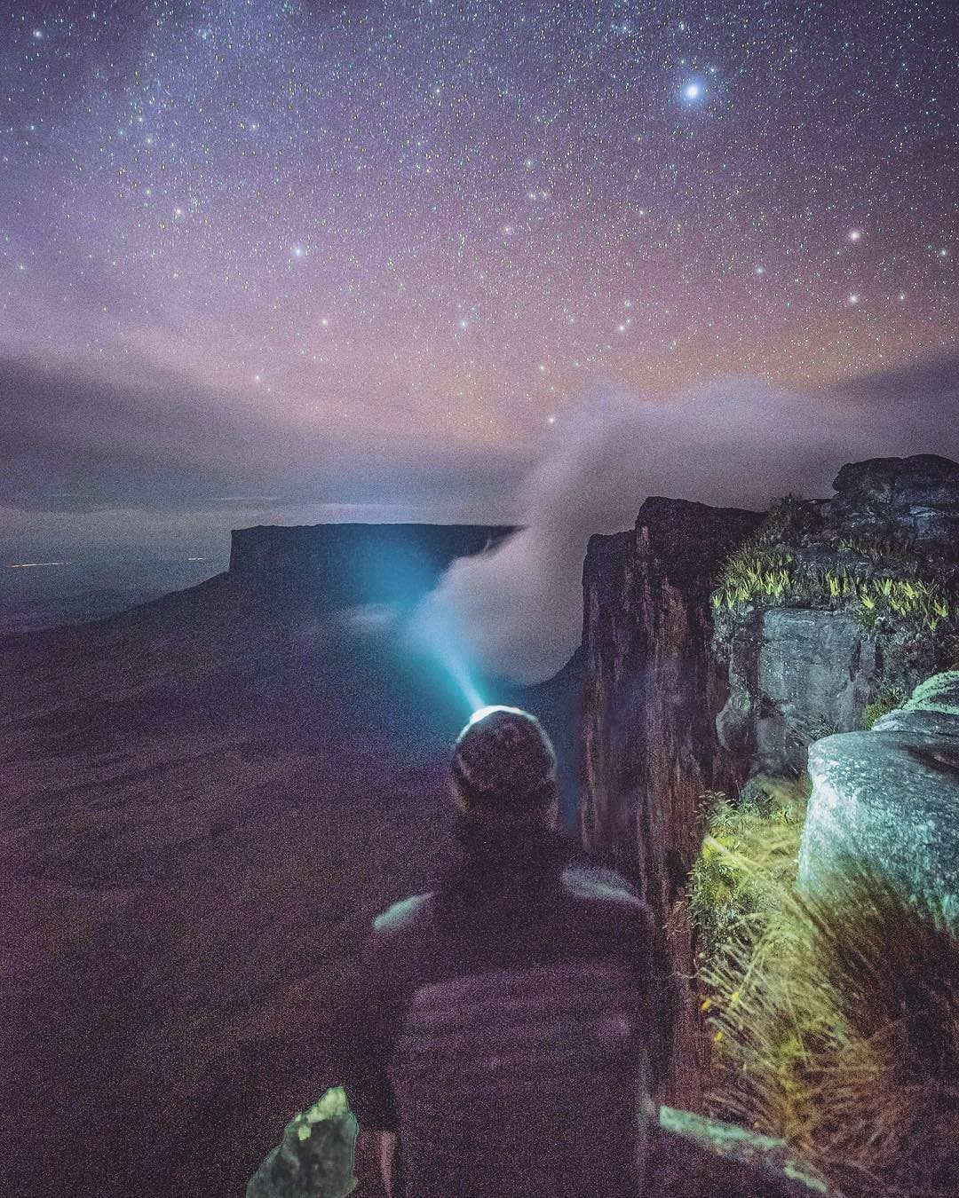 Up here, the stars feel like neighbors and the clouds are somewhere below your feet. A lone hiker with a headlamp stands on the rocky summit plateau of a tepui at night, gazing toward a flat-topped cliff shrouded in mist beneath a vast star-filled sky on the Mount Roraima hike