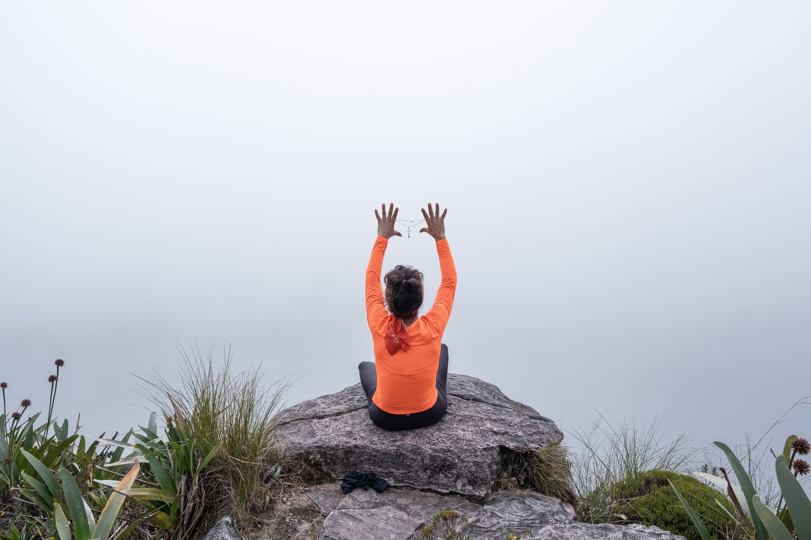 Hands up if the clouds came all the way to meet you. A person in an orange long-sleeve top sits cross-legged on a rocky summit outcrop, arms raised toward a dense white fog, surrounded by high-altitude bromeliads and grasses during the Mount Roraima hike