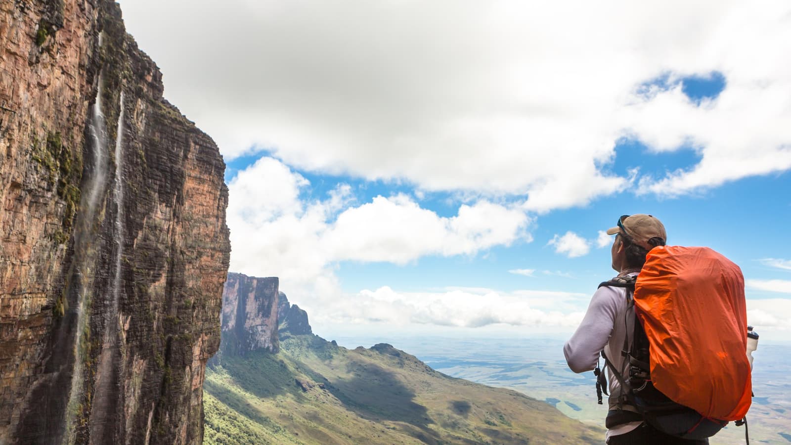 That moment when the cliff decides to show off and you just have to stop. A hiker with an orange pack gazes up at a sheer sandstone cliff with a thin waterfall cascading down it during the Mount Roraima hike, with a vast savanna plain stretching to the horizon below