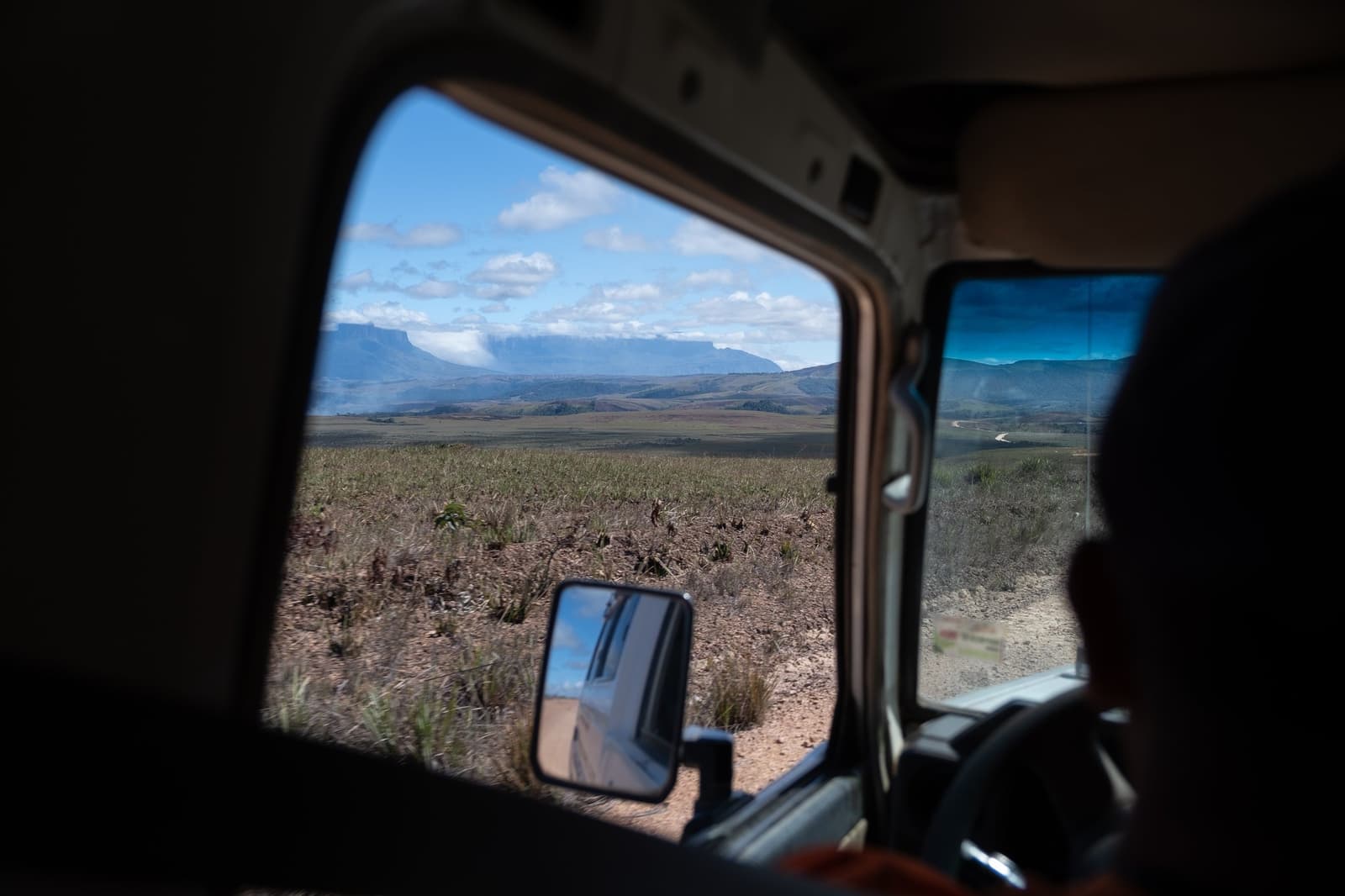 The tepui is right there, and yet the road keeps going. View through a 4x4 vehicle window of a vast open savannah with a flat-topped tepui mesa on the horizon during a Mount Roraima trek approach
