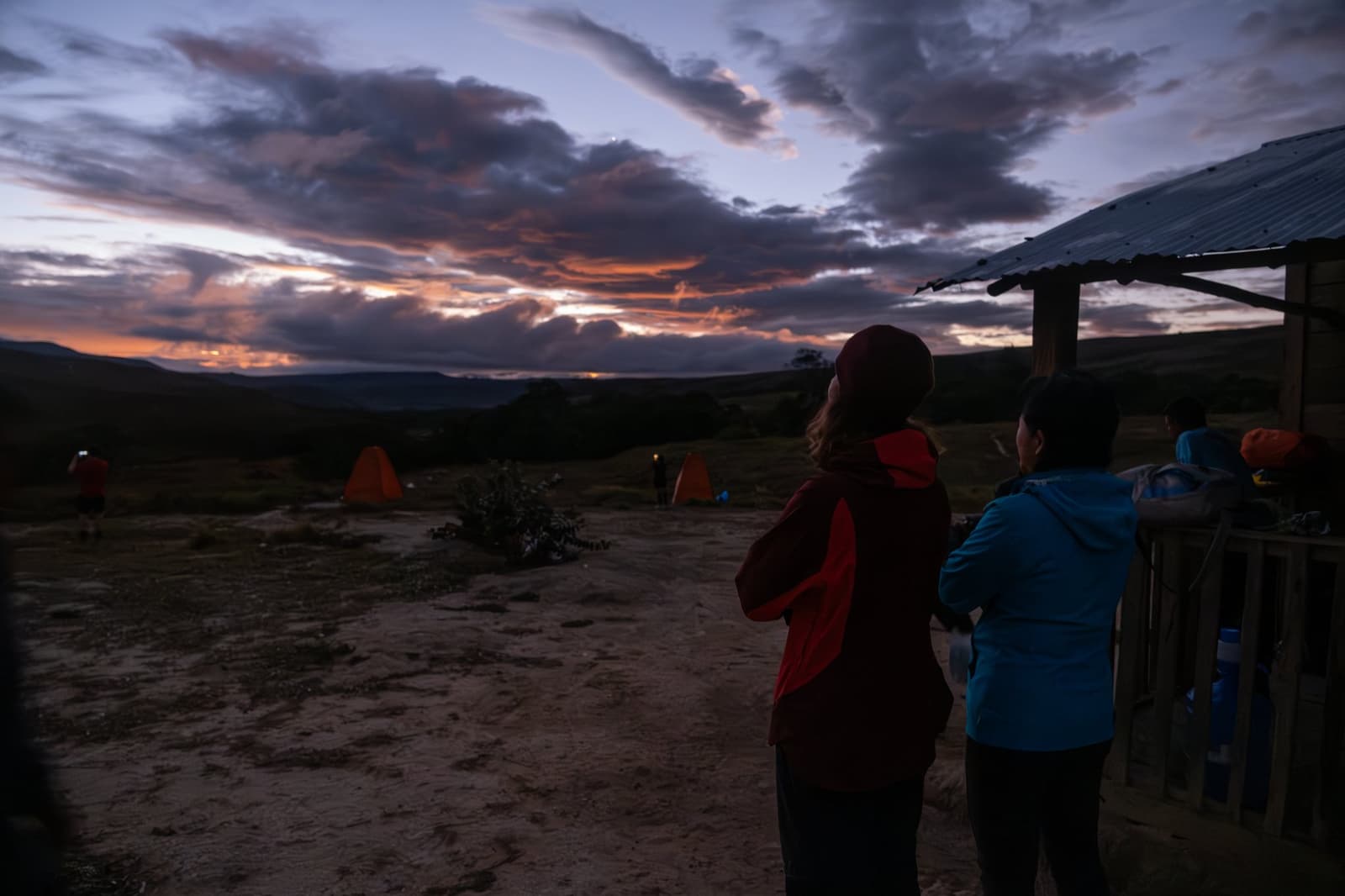 The end of a hard day's walking, and the sky puts on a show. Trekkers in warm jackets watch a dramatic orange and purple sunset from a hillside campsite during a Mount Roraima trek, with glowing orange tents visible in the background