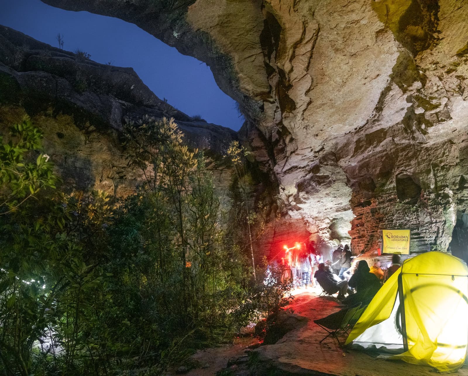 Your bedroom for the night: a 70-million-year-old sandstone ceiling, no reservation required. Trekkers gathered around a glowing campfire inside a vast sandstone rock shelter at night during a Mount Roraima trek, a yellow tent lit from within in the foreground and a Roraima Adventures banner visible on the cave wall