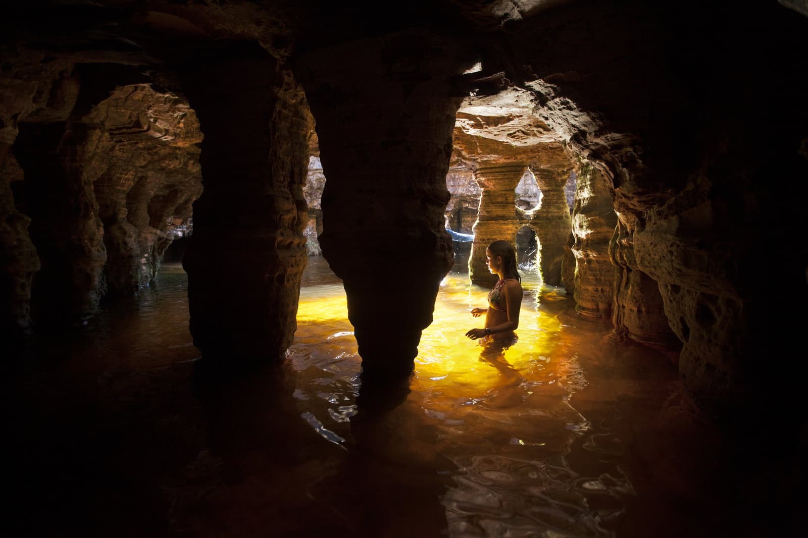 Light finds a way in, and so do you. A woman wades waist-deep through a glowing amber underground pool inside a sculpted sandstone cave on the Mount Roraima trek, light filtering through eroded rock arches