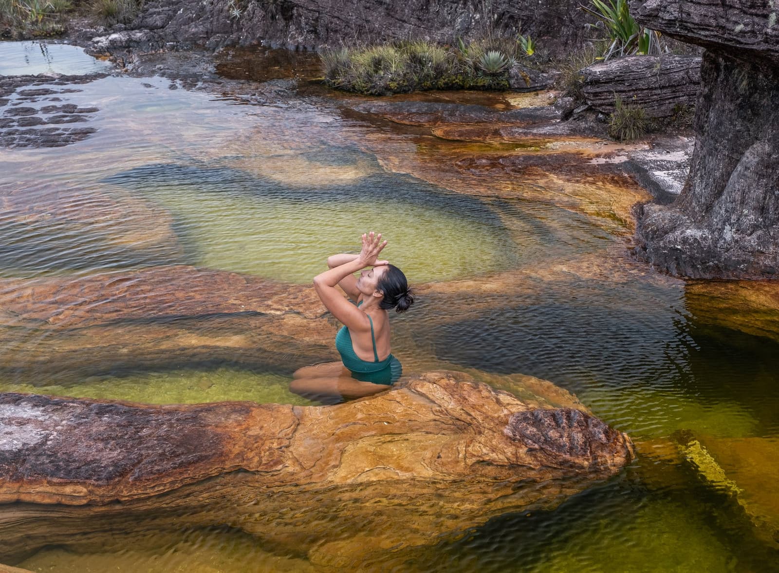 The summit has its own rules, and its own swimming holes. A woman in a teal swimsuit kneels in a shallow natural pool on the Mount Roraima trek, hands raised in a meditative pose above rust-coloured tepui rock carved by ancient water erosion