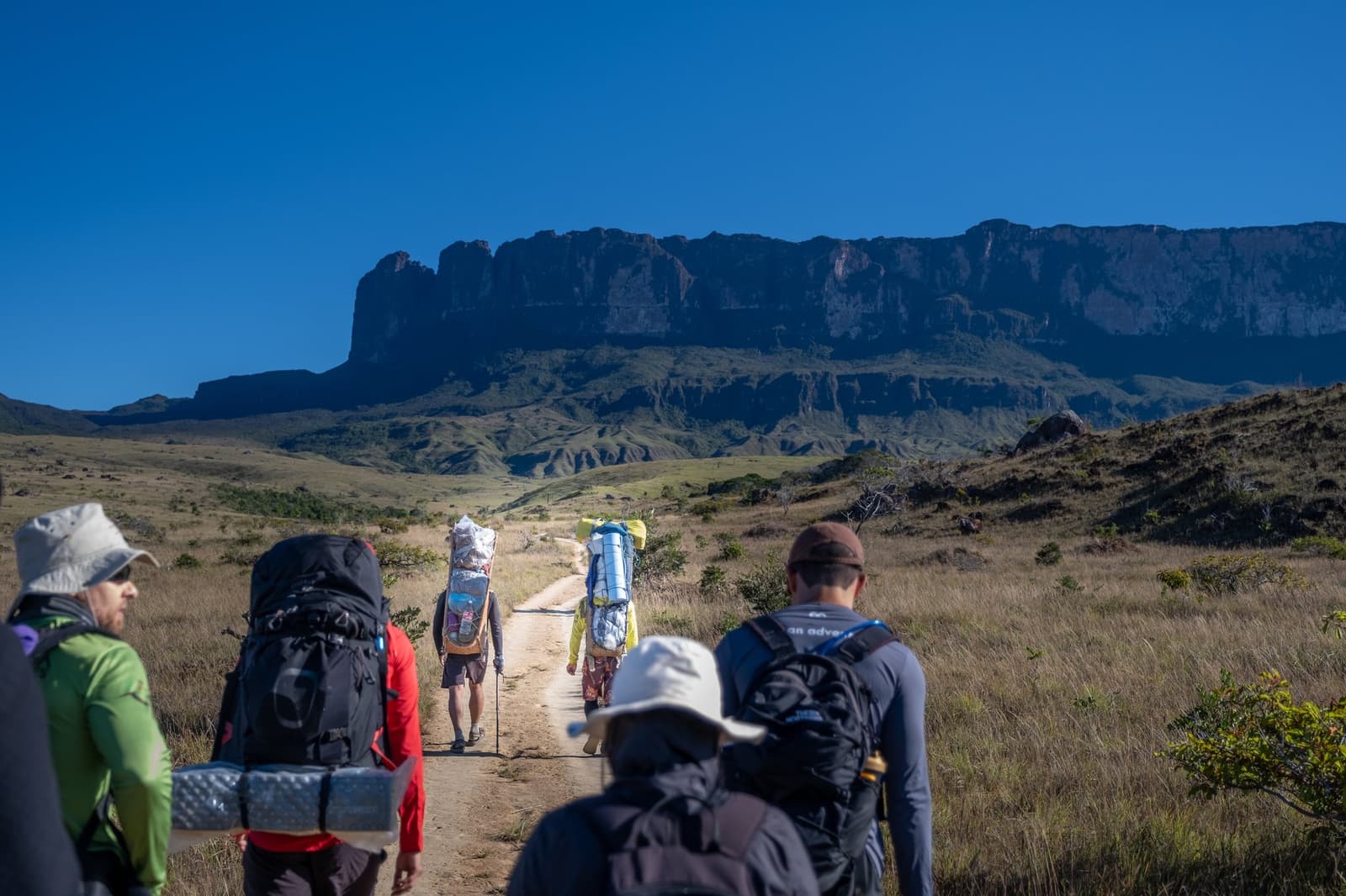 The mountain sets the pace. Everyone else just follows. A group of backpackers on the Mount Roraima trek walking a dirt trail toward the sheer sandstone cliffs of the tepui under a deep blue sky