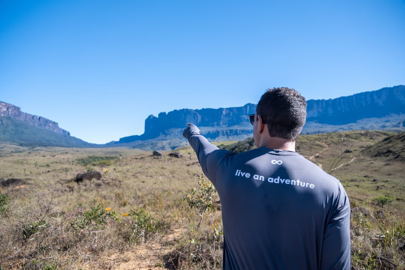That's your destination. No spoilers on what's waiting at the top. A guide in a 'live an adventure' shirt points toward the dramatic cliffs of a tepui on the Mount Roraima trek, with open savannah grassland stretching beneath a clear blue sky