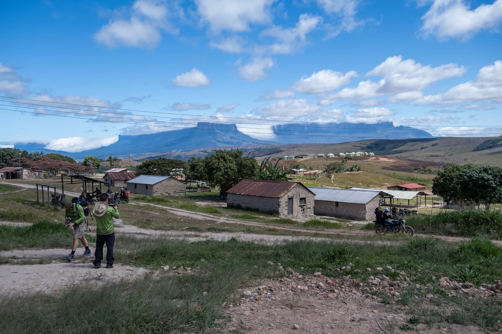 The village has a front-row seat to one of the world's great table mountains. Two photographers capture the view from a dirt path above a small village on the Mount Roraima trek, with a flat-topped tepui rising dramatically on the horizon under a blue sky with white clouds
