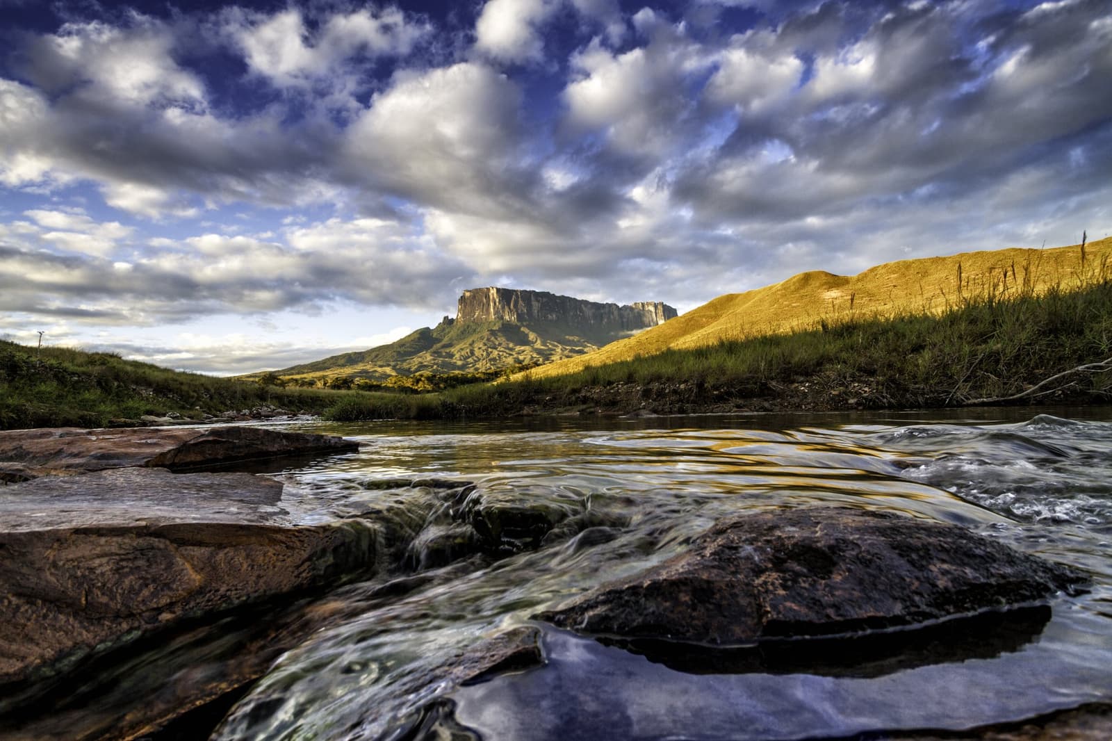 The river tells you you're getting close. Keep going. A shallow rocky river flows toward the flat-topped summit of Mount Roraima under a dramatic cloudscape on the Mount Roraima trek