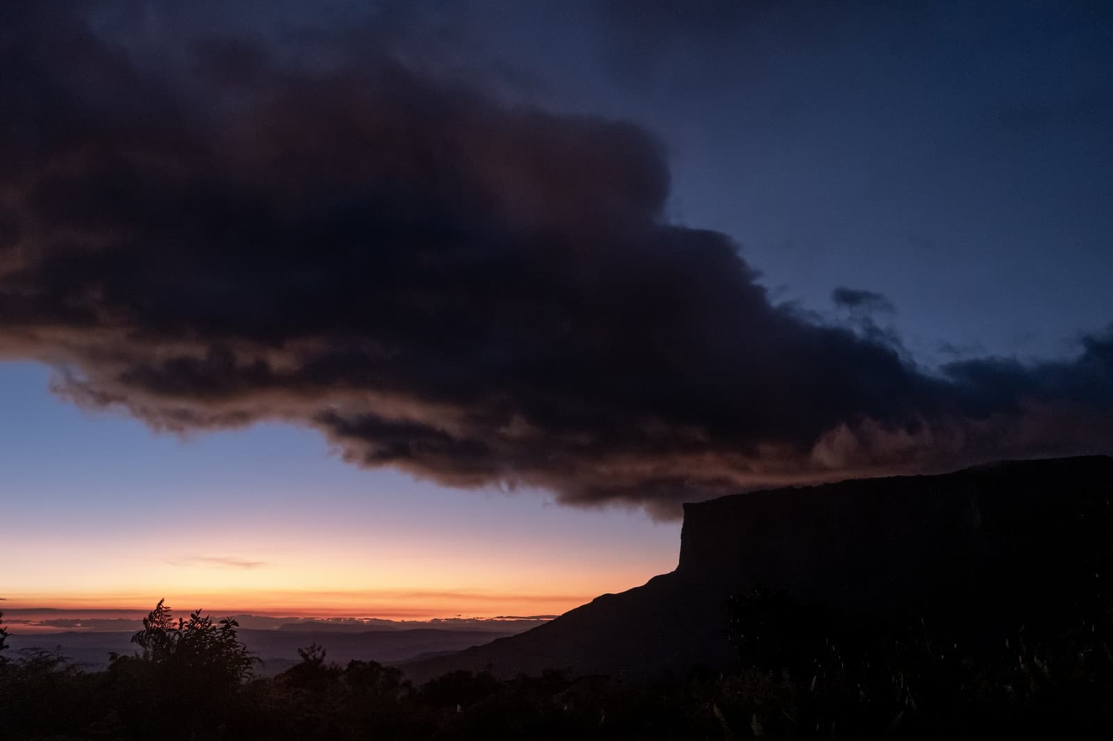 The mountain decides the mood, and tonight it chose dramatic. A dramatic storm cloud spreads over the silhouetted cliff edge of a tepui during a fiery Mount Roraima trek sunset, with an orange and deep-blue sky on the horizon