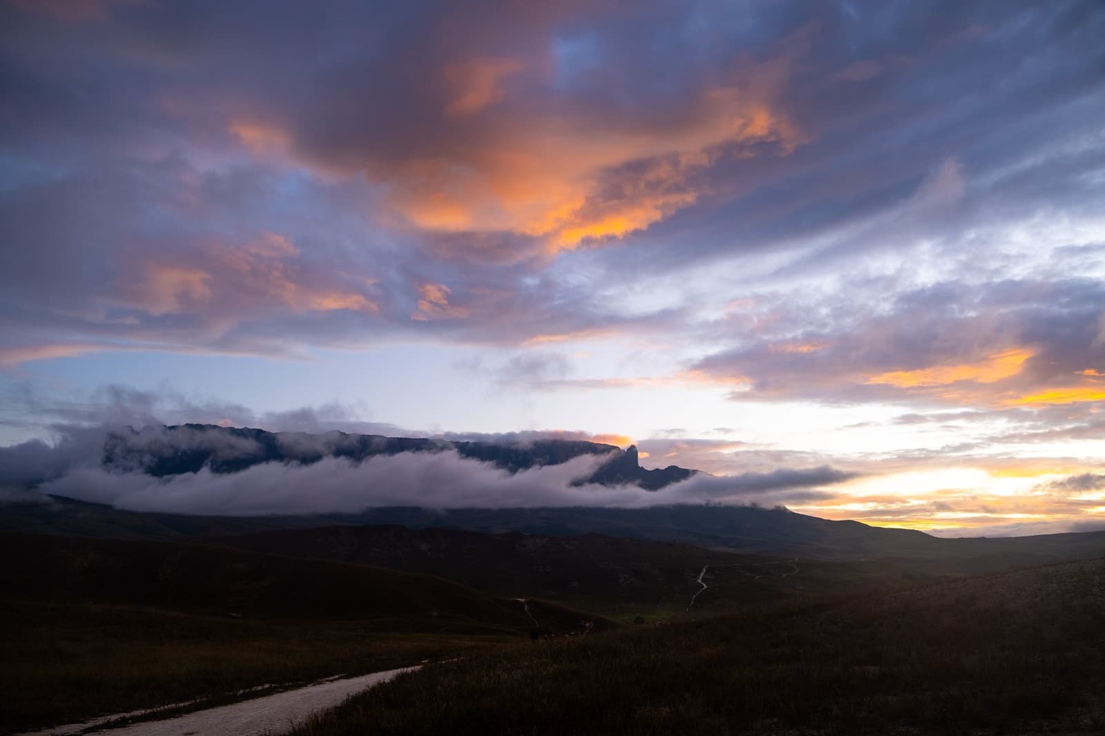 The light does something different up here every single evening. A dramatic sunset sky of orange and purple clouds hovers over a flat-topped tepui silhouette shrouded in low cloud during a Mount Roraima trek, with a winding trail visible across the dark savannah foreground