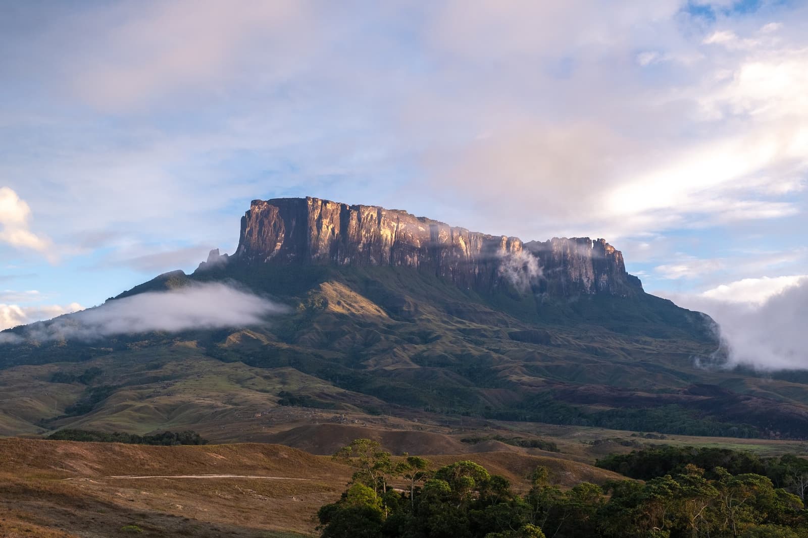 Ancient, flat, and completely indifferent to your sense of scale. The flat-topped sandstone massif of a tepui rises above rolling savannah grasslands during a Mount Roraima trek, with low clouds drifting around its sheer vertical cliffs in warm morning light
