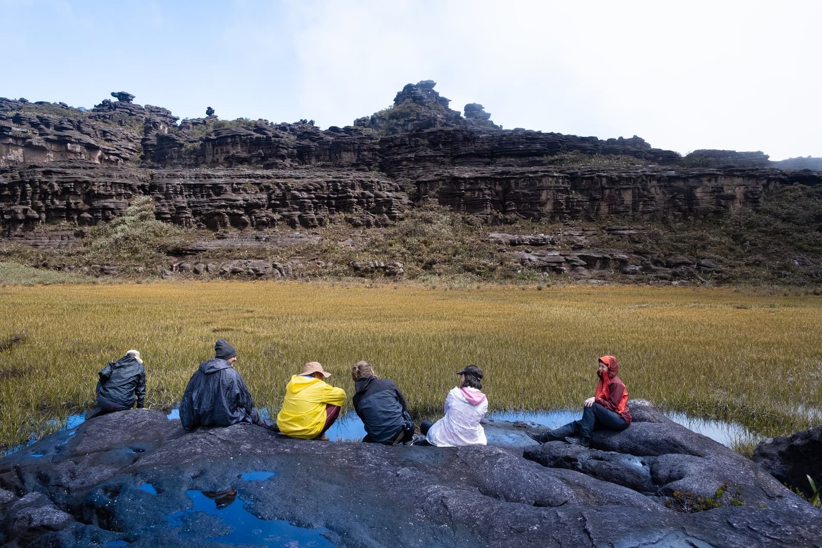 Sitting still on the summit plateau, letting the scale of it sink in. Six trekkers in rain gear sit on dark wet rocks overlooking a golden grassland meadow enclosed by ancient layered sandstone cliffs on the Mount Roraima trek