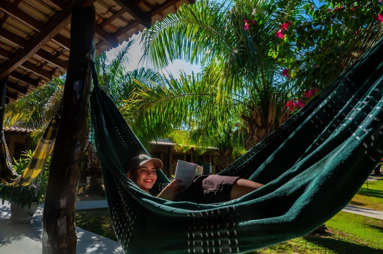 The book you never had time for back home? It fits perfectly here. A smiling traveller reads a book while lying in a green hammock beneath a shaded veranda, surrounded by palms and flowering bougainvillea at a Pantanal jaguar budget safari lodge