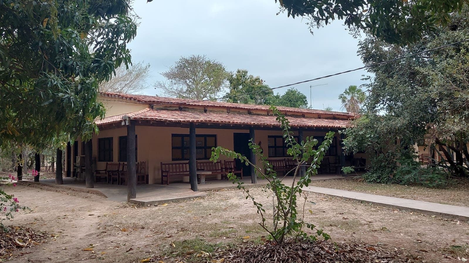 Simple, solid, and shaded, your base between game drives. A low-slung adobe lodge with a terracotta-tile roof and a shaded veranda lined with wooden benches, set among mature trees on a pantanal jaguar budget safari