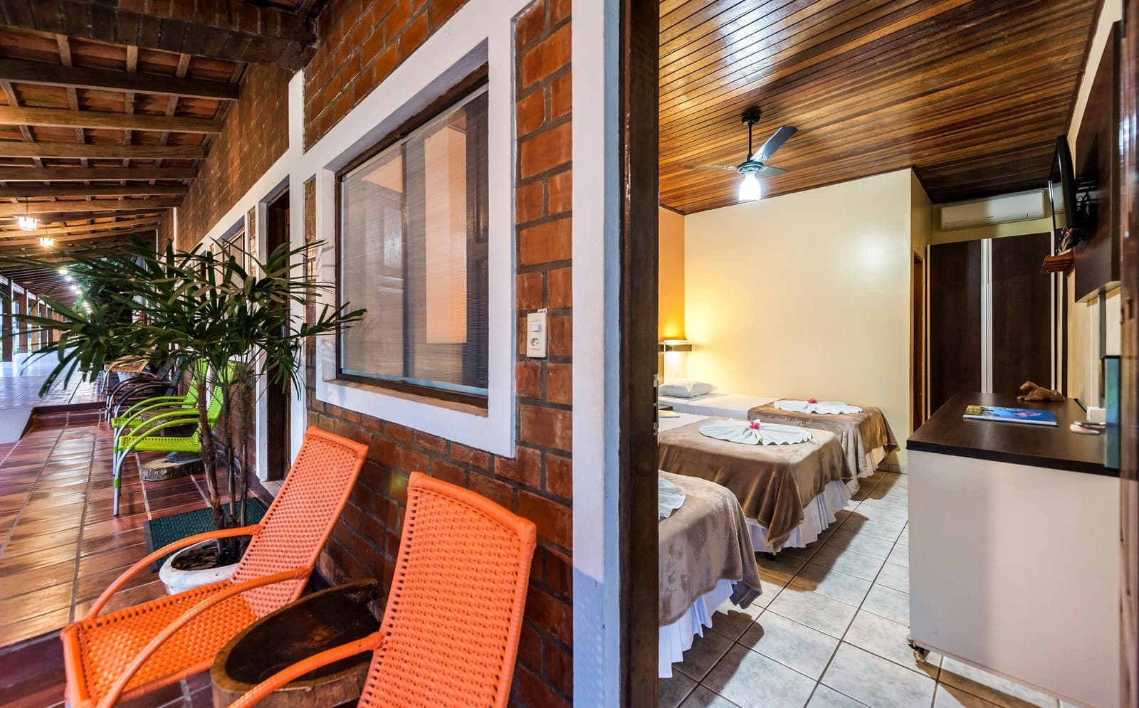 Simple, honest comfort after a long day on the water. Covered brick veranda with orange rattan chairs leading to a twin bedroom with ceiling fan and flower-decorated beds, typical of a pantanal jaguar budget safari lodge