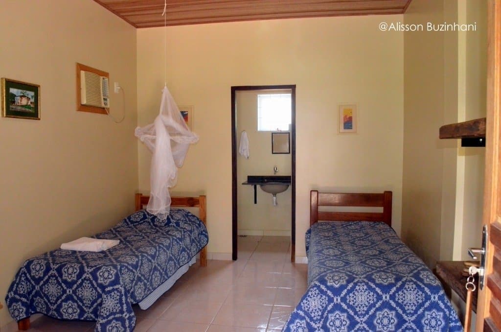 Simple, clean, and ready for an early wake-up call from the wildlife outside. Twin bedroom with blue patterned bedspreads, a hanging mosquito net, and an en-suite bathroom visible through the doorway at a simple Pantanal jaguar budget safari lodge