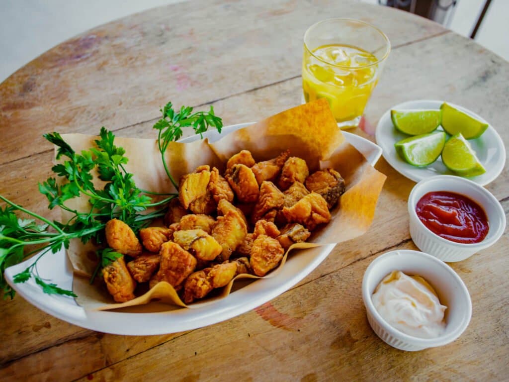 Cold drink, lime on the side, no decisions left to make. A platter of golden fried bites garnished with fresh parsley, served with lime wedges, ketchup, and cream sauce on a rustic wooden table during a pantanal jaguar safari porto jofre meal stop
