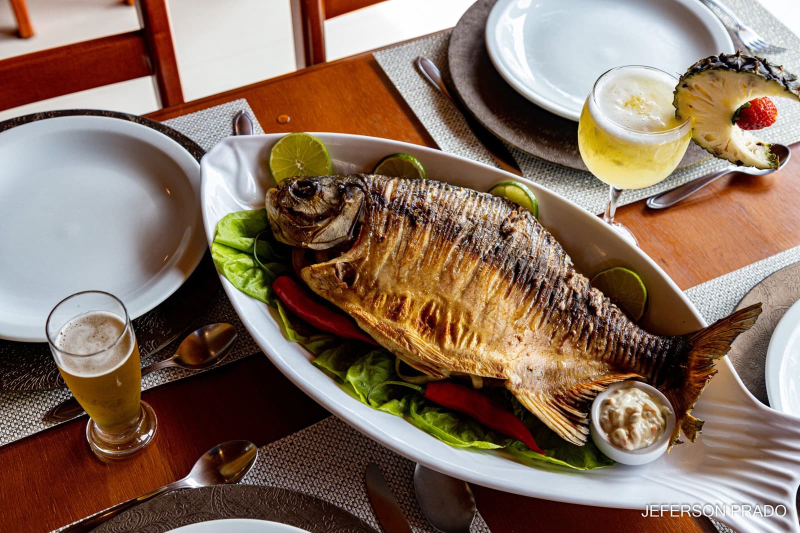 Fresh from the river, straight to the table, no argument needed. A whole grilled fish served on a white platter with lettuce, red peppers, lime slices, and a dipping sauce, alongside cold beer and a pineapple cocktail on a wooden dining table at a pantanal jaguar safari porto jofre lodge