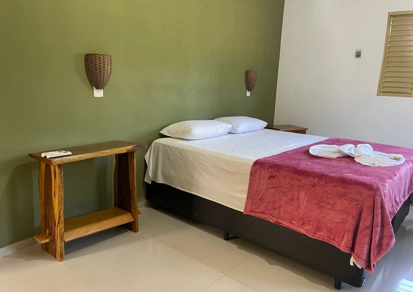 Simple, warm, and ready for you after a long day in the field. A neatly made double bed with a red blanket and folded towels in a simple lodge room with olive-green walls and rustic wooden furnishings, part of a pantanal jaguar safari porto jofre accommodation
