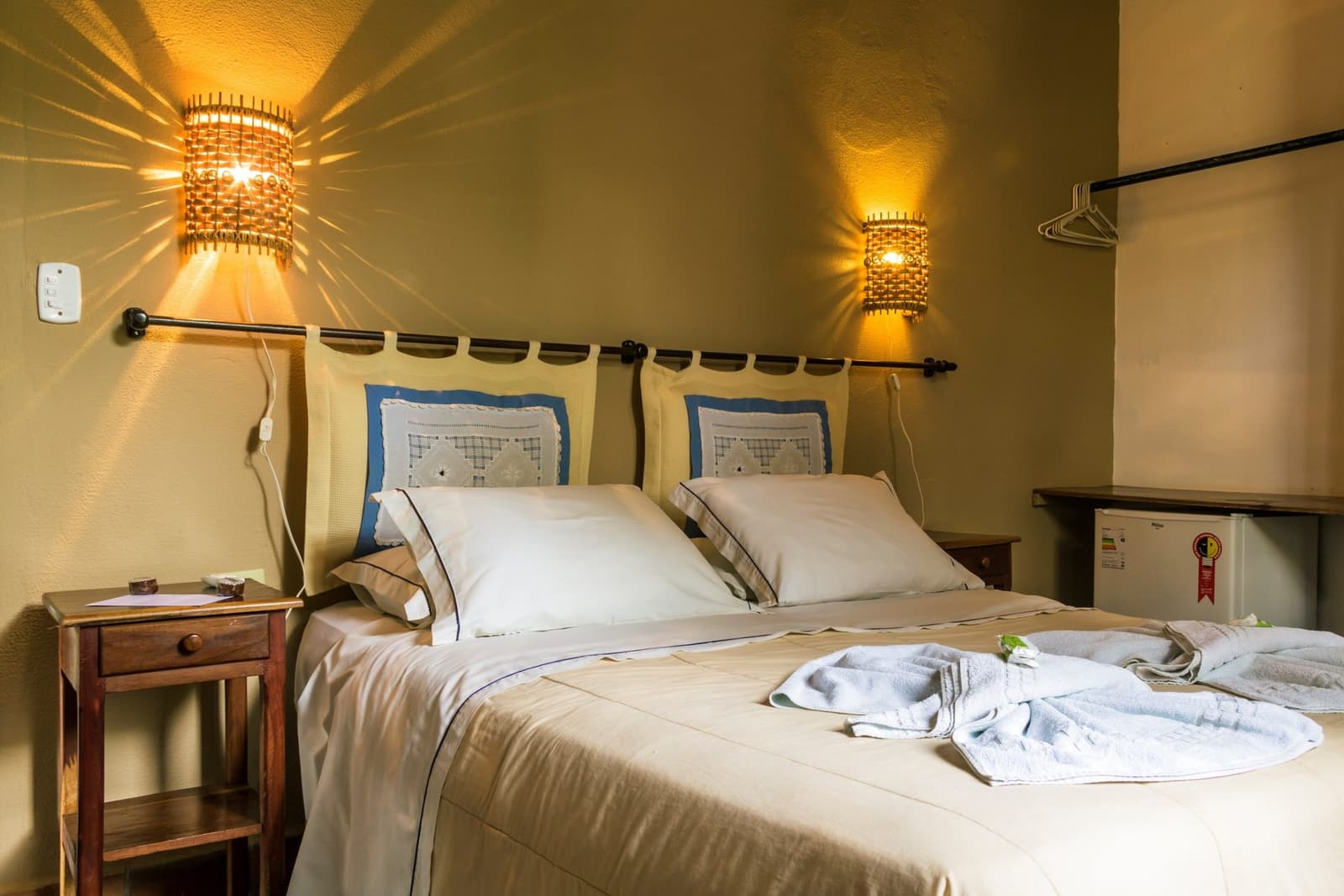 After a full day in the wetlands, this is exactly what you want to come back to. Cozy double room at a Pantanal tour from Campo Grande ecolodge, featuring warm woven wall sconces, cream bedding with decorative pillows, folded white towels and robes on the bed, a wooden nightstand, and a mini-fridge