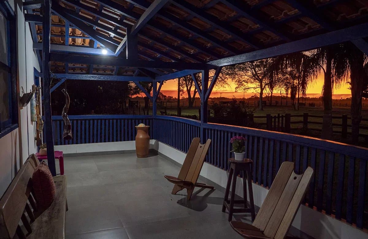 The best seat in the house costs nothing extra. A covered veranda with blue-painted wooden railings and rustic chairs overlooks a wide open landscape glowing with a deep orange sunset on a pantanal tour from campo grande
