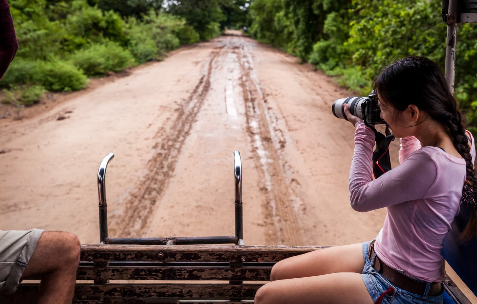 Mud on the road just means you're getting closer to the good stuff. A traveller shoots with a telephoto lens from the back of an open 4x4 vehicle on a red dirt road lined with green forest during a pantanal tour from campo grande