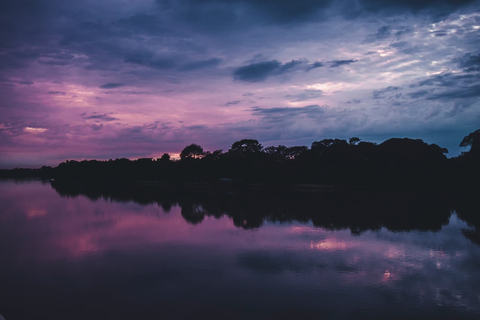 The Pantanal sky doesn't do subtle, even the river gets in on the show. A vivid purple and pink sunset sky reflected on a calm river, with a dark treeline silhouette along the horizon on a pantanal tour from campo grande