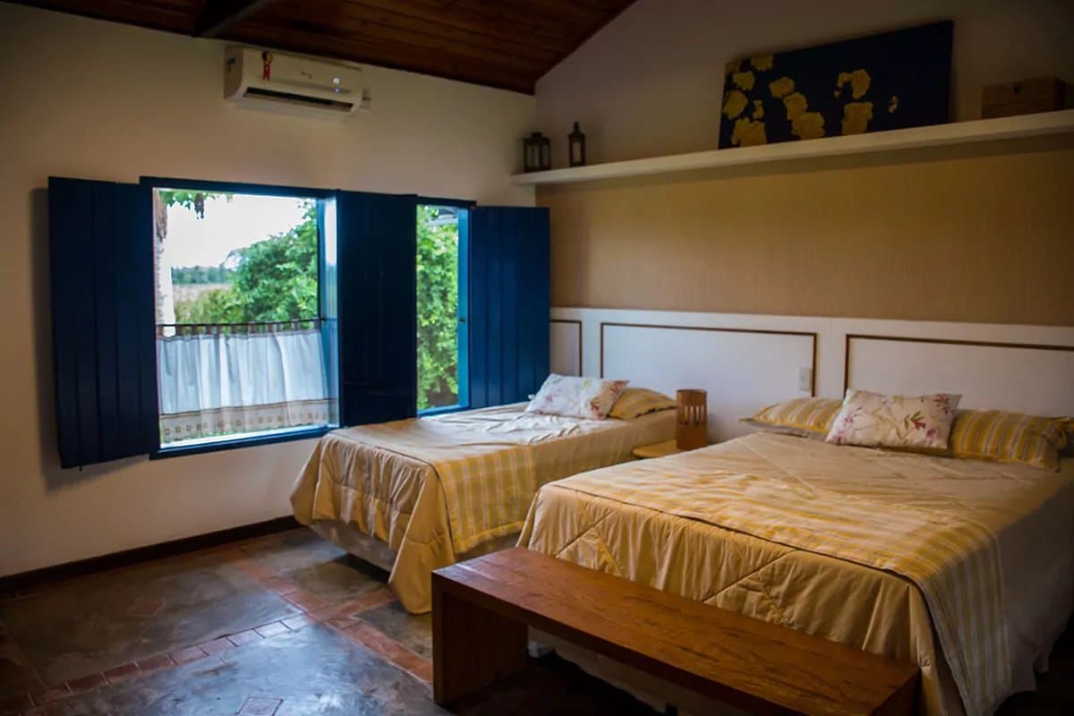 Your room has a view, and the jungle is doing its best to peek back in. A comfortable twin-bed lodge room with yellow-checked bedding, terracotta tile floor, wooden ceiling, and blue shuttered windows opening onto green vegetation, on a pantanal tour from campo grande