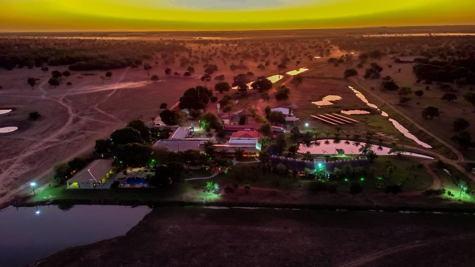 The lodge holds its own light long after the sun has other ideas. Aerial drone view of an illuminated Pantanal lodge at dusk, surrounded by wetland ponds and open savanna scrubland stretching to the horizon under a vivid yellow-orange sky — a base for a Pantanal Tour from Cuiabá