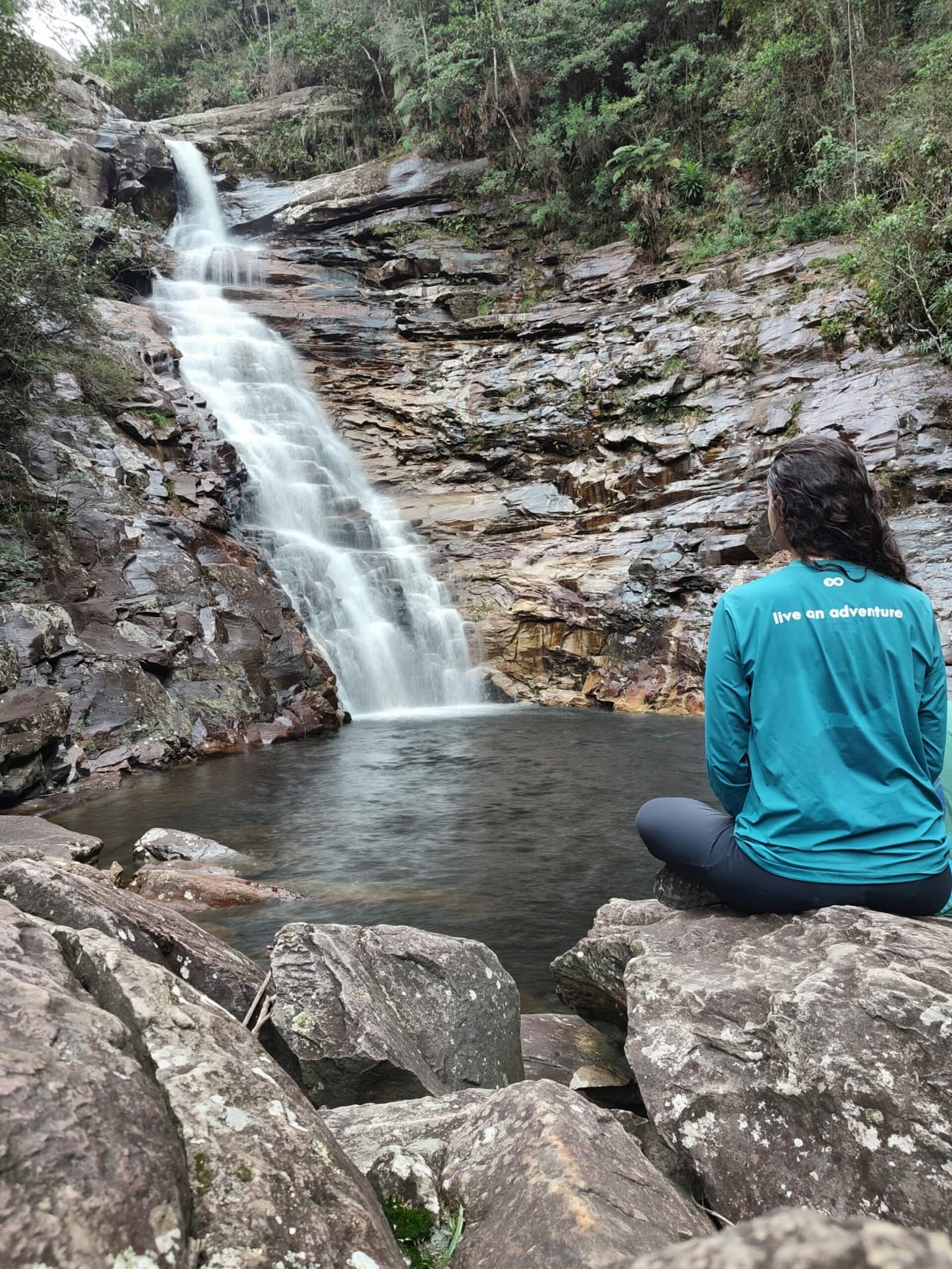The waterfall will still be there. Take a moment. A traveller in a teal top sits cross-legged on rocks during the Pati Valley hike, gazing at a silky waterfall cascading into a still pool surrounded by layered stone cliffs and forest
