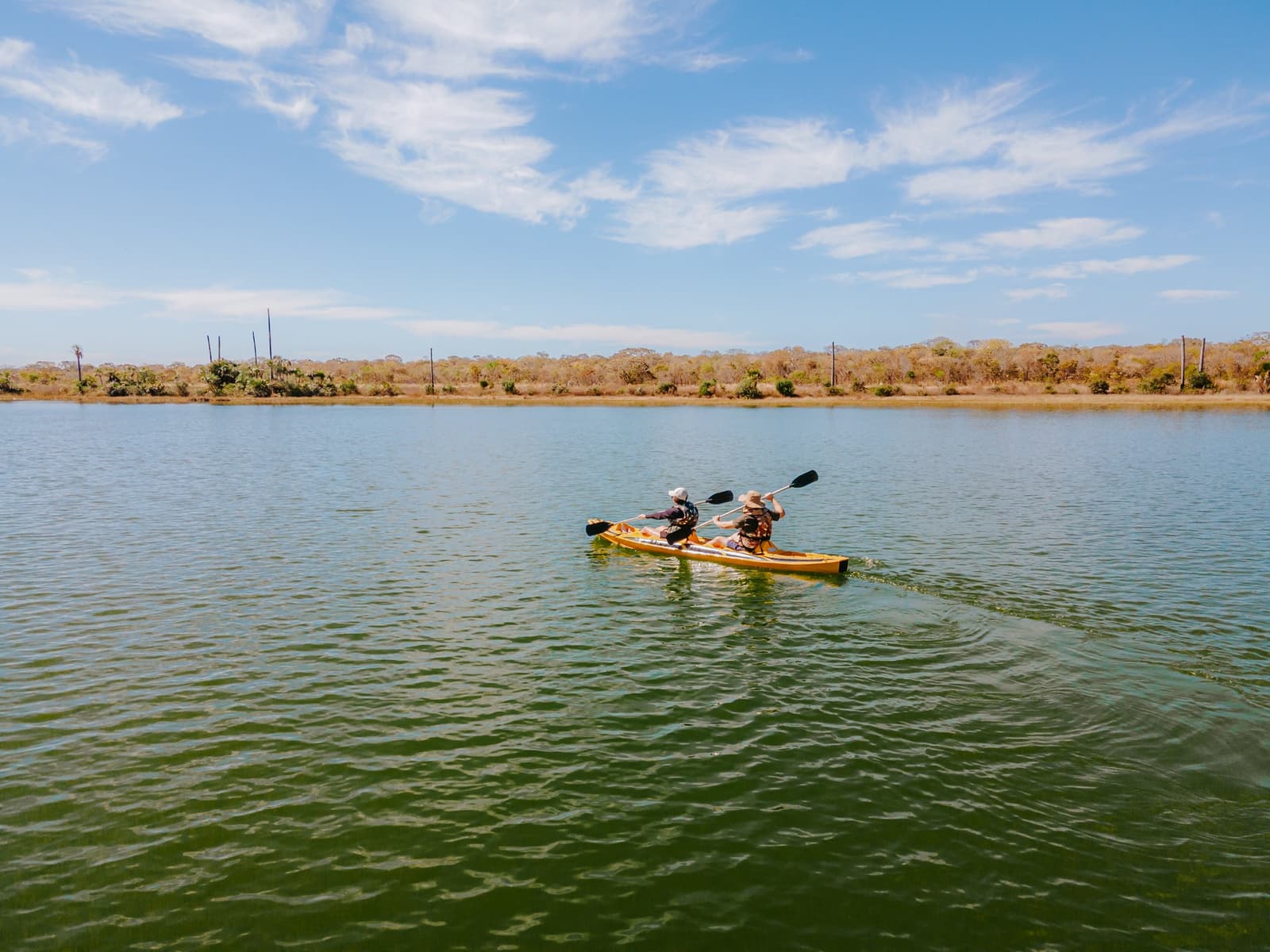 The Cerrado looks different at water level, quieter, and somehow bigger. Two paddlers in a yellow tandem kayak gliding across the calm green waters of Lagoa das Araras at Pousada Trijunção, with dry Cerrado woodland stretching along the far shore beneath a sunny blue sky