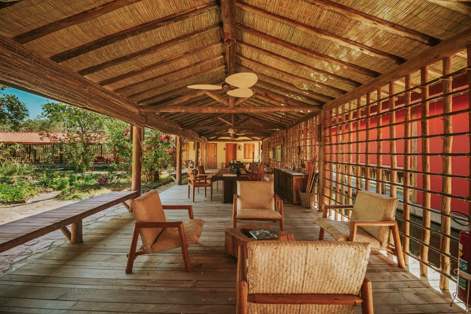 The kind of porch where trip stories get told and cold drinks disappear fast. Open-air lounge terrace at Pousada Trijunção with woven rattan chairs, rustic wooden deck, thatched palm roof and a lush garden view