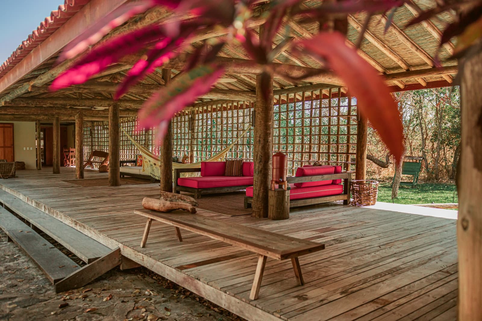 The kind of veranda where afternoons have a way of disappearing. Open-air lounge at Pousada Trijunção featuring a rustic thatched roof supported by log columns, red-cushioned wooden sofas, hanging hammocks, and a wide timber deck framed by pink bougainvillea